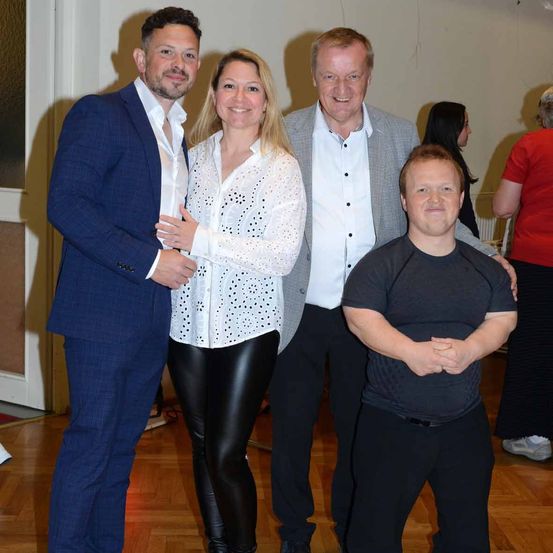 Vier Menschen stehen zusammen in einem Raum. Der Mann auf der linken Seite lächelt und hat seine Hand auf der Schulter der Frau in der Mitte. Die Frau hat ihre Hand auf der Brust des Mannes. Der Mann auf der rechten Seite schaut in die Kamera.