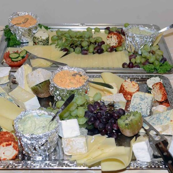 Zwei Tabletts mit Käse und Obst stehen auf einem Tisch. Das obere Tablett hat verschiedene Käsesorten, Trauben und eine Scheibe Kiwi. Das untere Tablett hat mehrere Käsesorten, Trauben und einen Löffel.