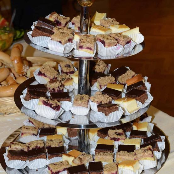 Ein dreistöckiger Kuchenständer mit einer Auswahl an Desserts, darunter Schokoladen- und Beeren-Toppings, die in Cupcake-Förmchen angeordnet sind. Im Hintergrund befinden sich ein Korb mit Brot und eine Schale mit Obst.
