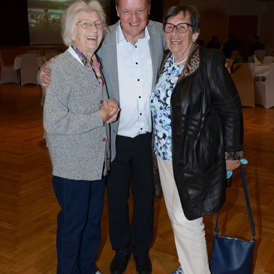 Drei ältere Personen stehen lächelnd zusammen und posieren für das Foto, der Hintergrund mit Tischen und Stühlen ist unscharf.