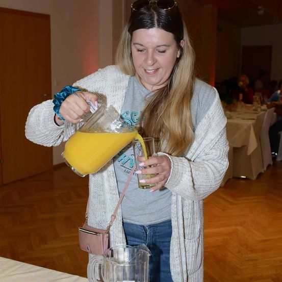 Eine Frau gießt Orangensaft aus einem Krug in ein Glas und lächelt bei einer gesellschaftlichen Veranstaltung.
