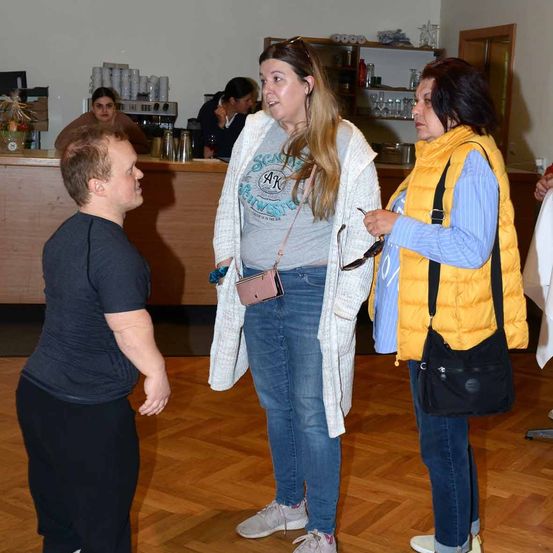 Drei Personen stehen in einem Raum, zwei Frauen und ein Mann. Die Frau in der Mitte trägt einen weißen Cardigan. Eine andere Frau trägt eine gelbe Weste. Der Mann trägt ein schwarzes T-Shirt. Hinter ihnen steht eine Theke mit einer Kaffeemaschine.