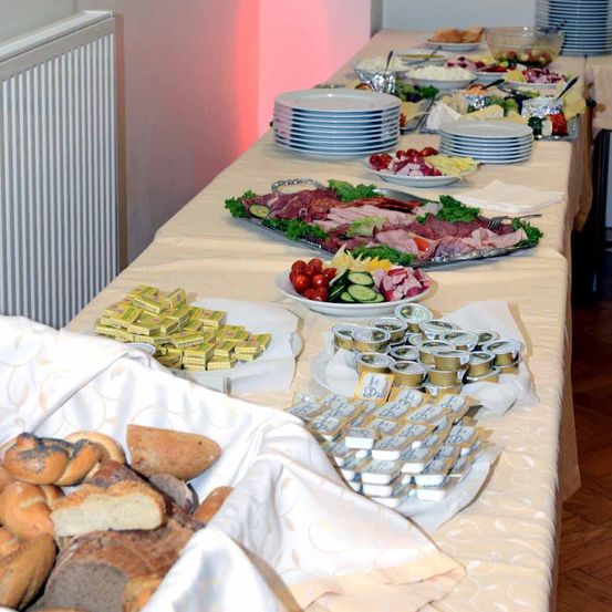 Ein langer Tisch mit einer weißen Tischdecke zeigt eine Vielzahl von Speisen, darunter Brot, Fleisch, Gemüse und Käse. Schüsseln mit Obst und Salaten sind ebenfalls vorhanden.