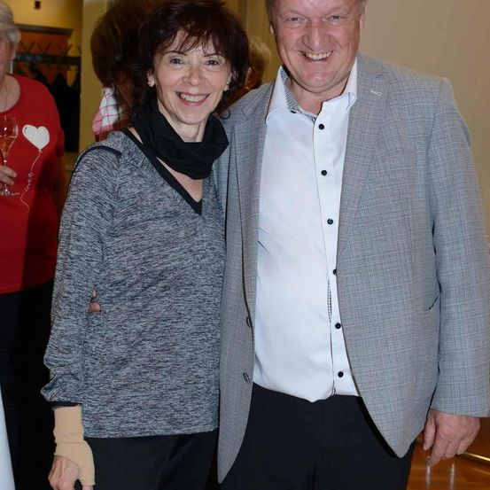 Ein lächelndes Paar, eine ältere Frau in einem grauen Top und ein Mann in einem grauen Blazer, stehen bei einer Veranstaltung drinnen zusammen, mit einer Person, die einen Glas Wein im Hintergrund hält.