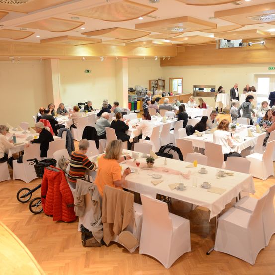 Ein Speisesaal mit mehreren Tischen, die mit weißen Tüchern bedeckt sind, beherbergt eine Veranstaltung mit vielen Teilnehmern. Der Saal hat Holzböden, weiße Wände und eine hohe Decke mit Deckenleuchten.