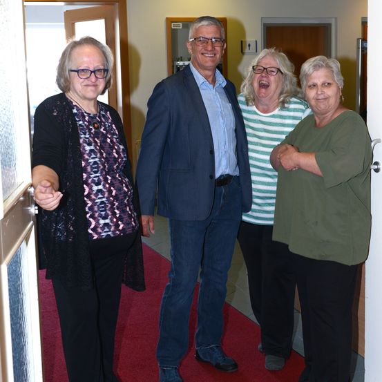 Vier Personen, zwei Frauen und zwei Männer, stehen lächelnd auf einem roten Teppich vor einer offenen Tür, was auf eine feierliche Veranstaltung hindeutet.