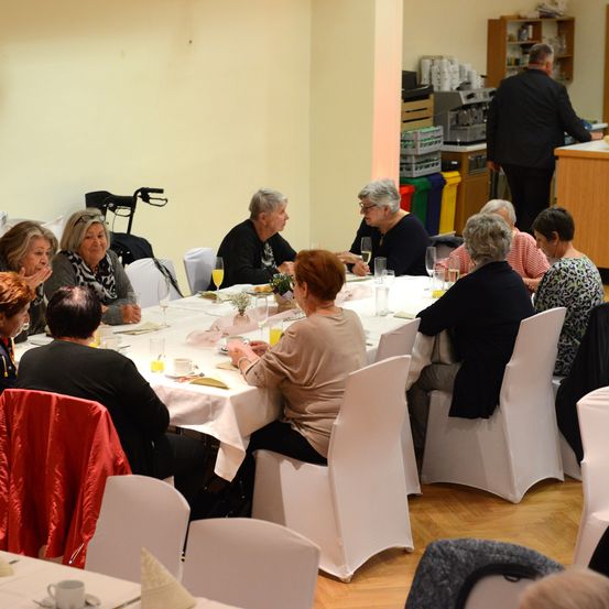 Eine Gruppe älterer Menschen sitzt um einen langen Tisch mit weißen Tischdecken, Tassen und Weingläsern. Eine Person steht hinter der Theke und bereitet möglicherweise Kaffee zu.