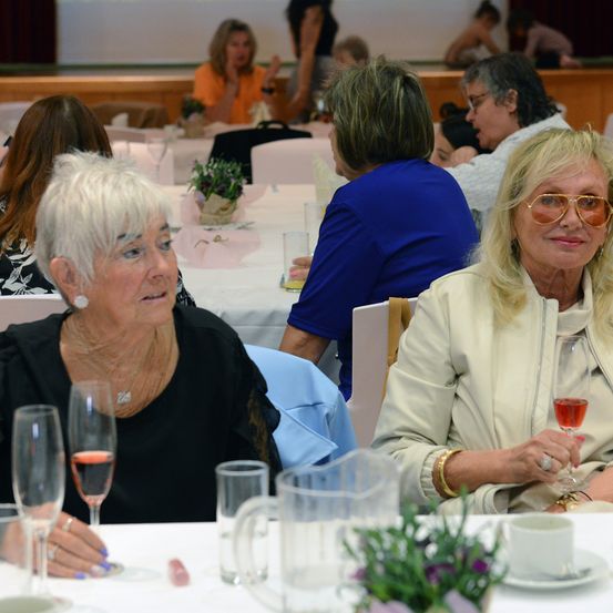 Eine Gruppe von Frauen versammelt sich an einem Tisch für eine Veranstaltung. Zwei ältere Frauen sitzen vorne, lächeln und halten Weingläser in der Hand. Ein Krug und Gläser stehen auf dem Tisch.
