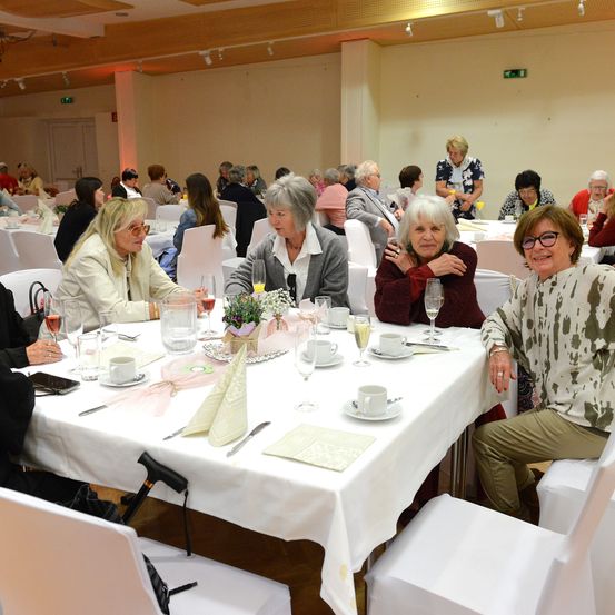 Mehrere ältere Frauen und Männer sitzen an einem großen Tisch in einem Bankettsaal, lächeln und plaudern, mit Weinläsern, Tassen und Tellern auf dem Tisch.