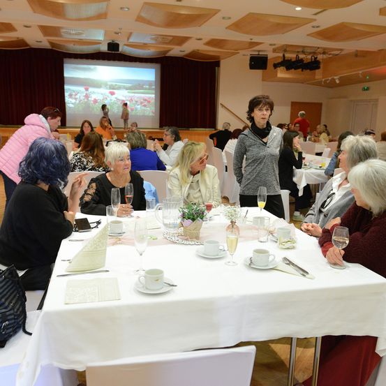Eine Indoor-Veranstaltung mit mehreren Frauen, die um einen Tisch sitzen, mit Tassen, Gläsern und Blumen. Ein Beamerschirm im Hintergrund mit Leuten darauf.