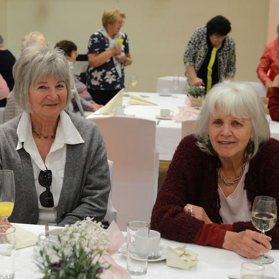 Zwei ältere Frauen sitzen an einem Tisch mit Weinläsern, eine trägt einen grauen Pullover und die andere eine rote Strickjacke. Hinter ihnen sitzen mehrere andere Personen, und eine Frau steht in der Nähe eines Tisches mit einer Blumenarrangement.