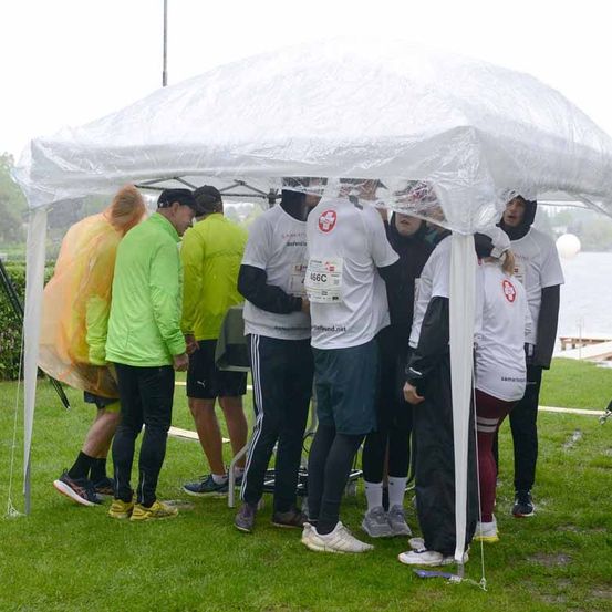 Eine Gruppe von Läufern in Regenkleidung steht unter einem durchsichtigen Zelt, einer trägt ein weißes Shirt mit der Nummer 456C. In der Nähe sind Menschen in Regenmänteln und Turnschuhen.