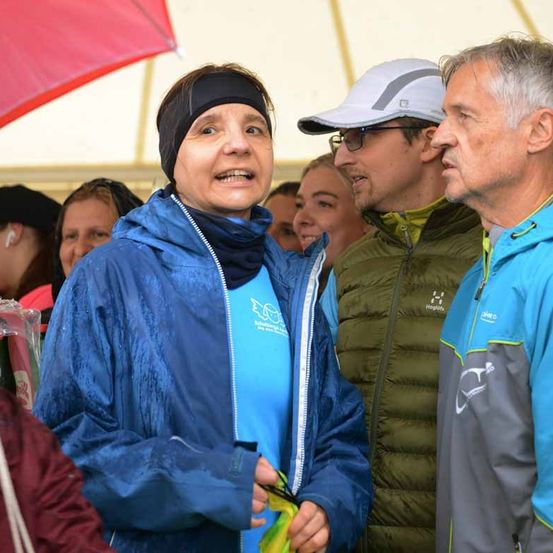 Eine Frau in einer blauen Jacke und einem Stirnband spricht mit zwei Männern in einem überfüllten Raum mit einem Regenschirm und einem Zelt im Hintergrund.