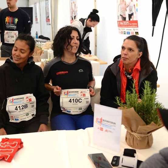 Drei Frauen sitzen an einem Tisch, zwei halten Startnummern. Sie lächeln, möglicherweise bei einer Marathonveranstaltung. Ein Topf mit Pflanze und ein Banner stehen auf dem Tisch.