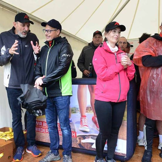 Mehrere Personen stehen in einem Zelt mit einem Banner, das Schuhe zeigt. Eine Frau in einer rosa Jacke hält einen Becher. Ein Mann in einer schwarzen Jacke spricht mit einem anderen Mann. Hinter ihnen hält ein Mann eine Flasche. In der Nähe steht eine Kiste mit Bananen.