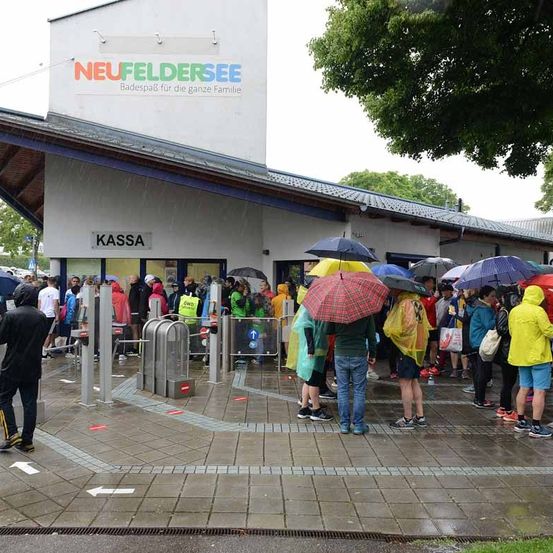 Eine Menschenmenge mit Regenschirmen steht in der Schlange am Eingang des Neufeldersees, einem familienfreundlichen Schwimmbereich.