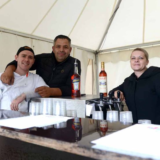 Drei Personen stehen hinter einem Bartresen in einem Zelt und lächeln für ein Foto. Ein Mann auf der linken Seite trägt ein weißes Kochoutfit, während der Mann in der Mitte eine schwarze Jacke trägt. Eine Frau auf der rechten Seite trägt eine schwarze Jacke. Flaschen von Aperol und Lillet stehen auf dem Tresen.