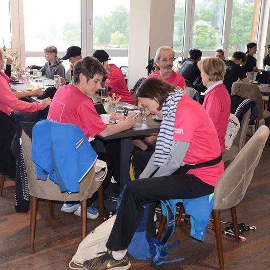 Eine Gruppe von Menschen sitzt an Tischen in einem Restaurant. Sie tragen rosa Hemden. Eine Frau sitzt auf dem Boden und hält ein Stück Papier. Die Stühle sind um die Tische herum angeordnet. Der Boden ist aus Holz.