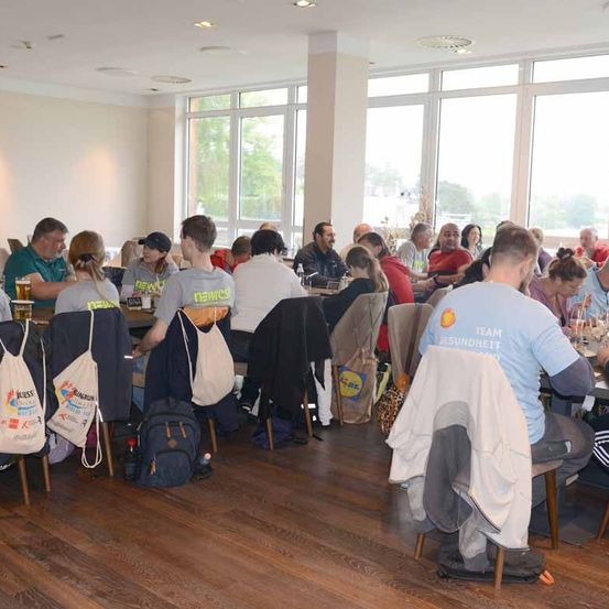 Eine Gruppe von Menschen sitzt an Tischen in einem Raum mit großen Fenstern. Einige tragen T-Shirts mit verschiedenen Logos, und es gibt Rucksäcke und Taschen auf Stühlen. Gläser mit Getränken stehen auf den Tischen.