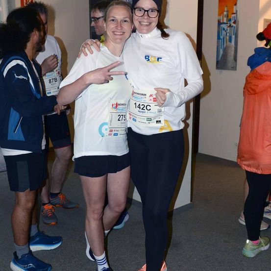 Zwei Frauen, eine mit Brille, posieren für ein Foto, während andere hinter ihnen stehen, wahrscheinlich bei einem Marathon-Event. Sie tragen nummerierte Shirts, und ein Banner hängt an der Wand.