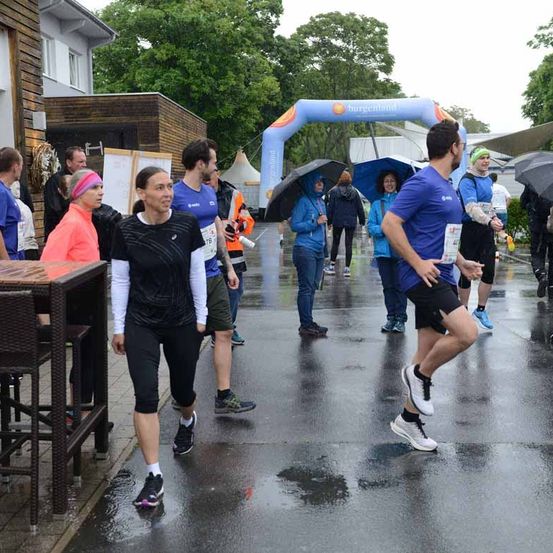 Eine Gruppe von Menschen nimmt an einem Laufevent an einem regnerischen Tag teil, trägt Laufausrüstung und hält Regenschirme. Das Event ist durch einen blauen Bogen gekennzeichnet.
