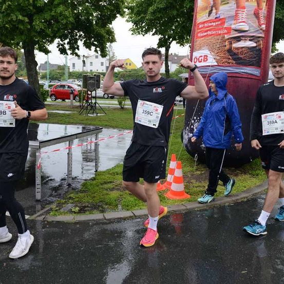 Läufer nehmen an einem Marathon teil. Der Mann in der Mitte trägt eine Uhr und hat seine Fäuste erhoben. Rechts geht eine Frau in einer blauen Regenjacke. Bäume und geparkte Autos sind im Hintergrund.