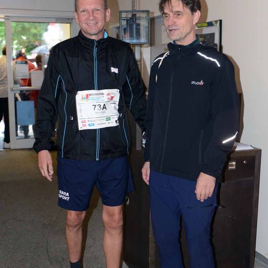 Zwei Männer stehen in einem Korridor, beide tragen schwarze Jacken mit weißen Texten. Der Mann auf der linken Seite hat '73A' auf seiner Jacke, während der Mann auf der rechten Seite 'IFLOV' hat. Hinter ihnen befindet sich eine Glastür mit einem Flur und einer Glasvitrine.
