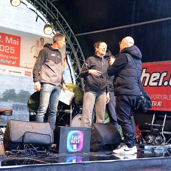 Drei Personen stehen auf einer Bühne unter einem großen Banner. Ein Mann hält ein Papier, eine Frau lacht und ein weiterer Mann spricht zu ihnen. Musikequipment ist im Hintergrund zu sehen.