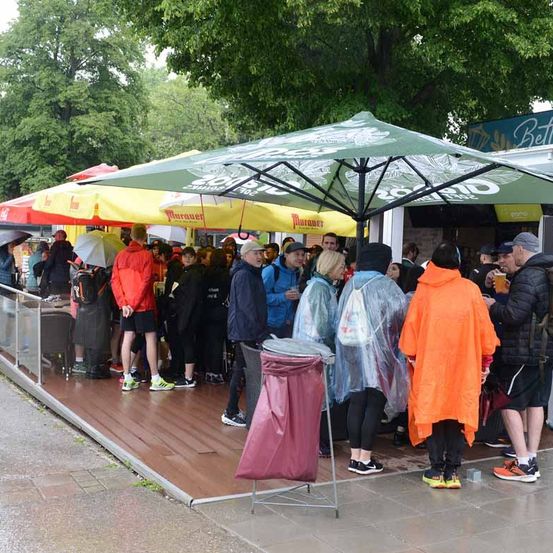 Menschen in Regenmänteln stehen unter Regenschirmen in einer Schlange auf einem nassen Bürgersteig. Bäume und ein Gebäude mit einem Schild sind im Hintergrund.