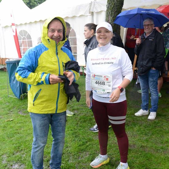 Ein Mann und eine Frau stehen auf einem Rasenfeld und tragen Laufausrüstung. Dahinter hält eine Person einen Schirm, und ein Mann in einem schwarzen Mantel ist zu sehen. Sie befinden sich wahrscheinlich bei einem Marathonevent.
