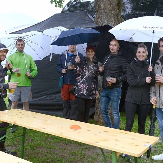 Eine Gruppe von Menschen lächelt und posiert für ein Foto, während sie Regenschirme und Getränke in einem Park halten. Sie stehen vor einer Holzbank.