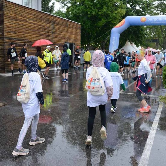 Eine Gruppe von Menschen geht im Regen, trägt Regenmäntel und Sneakers. Einige haben Regenschirme dabei. Hinter ihnen ist ein Gebäude und ein blauer Bogen zu sehen. Bäume und Zelte sind im Hintergrund.