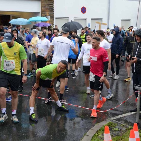 Marathonteilnehmer versammeln sich auf einer nassen Fläche. Einige tragen Startnummern, einer lehnt sich vor. Regenschirme und Jacken sind zu sehen. Gebäude, ein rotes Schild und Verkehrskegel im Hintergrund.