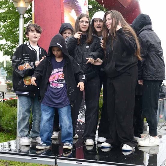 Eine Gruppe von Kindern und Jugendlichen steht auf einer nassen Bühne, wahrscheinlich in einem Park. Sie tragen alle schwarze Jacken und Hosen. Der Junge in der Mitte trägt eine Kapuzenjacke mit einem rosa Design. Sie posieren für ein Foto.