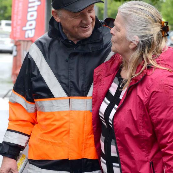 Ein älterer Mann in einer orangefarbenen und schwarzen reflektierenden Jacke lächelt, während er eine ältere Frau in einer rosa Jacke ansieht.