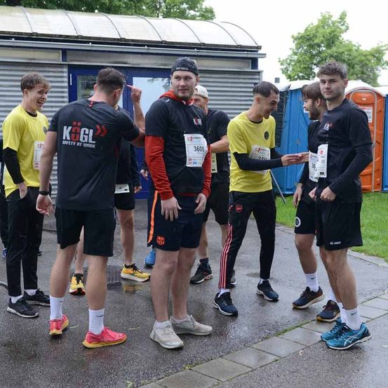 Mehrere Männer stehen auf einem nassen Bürgersteig, wahrscheinlich vor einem Marathon. Sie tragen nummerierte Startnummern und einige tragen Sportbekleidung. Im Hintergrund sind mobile Toiletten zu sehen.