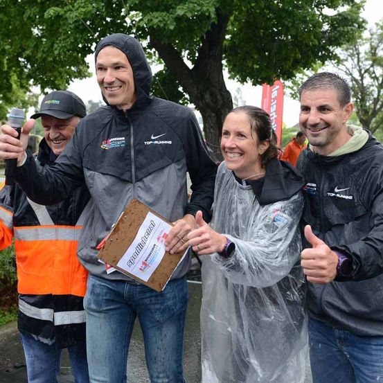 Vier Menschen stehen im Regen; einer hält ein Mikrofon und klatscht. Zwei Männer in Jacken, einer trägt eine orange Weste, lächeln. Eine Frau in einer Plastikregenjacke lächelt mit erhobenem Daumen.