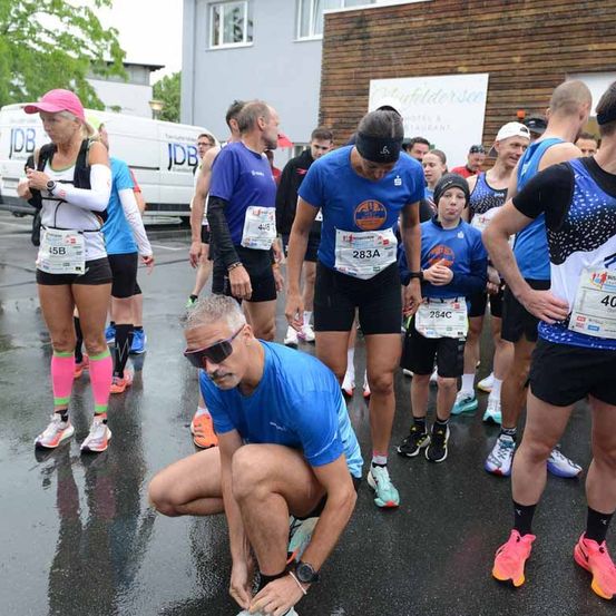 Läufer mit verschiedenen farbigen Startnummern stehen auf einer nassen Straße in einer Reihe. Ein Mann in einem blauen Shirt schnürt seine Schuhe. Im Hintergrund ist ein Gebäude mit einem Schild, das Stiddersee liest, zu sehen.