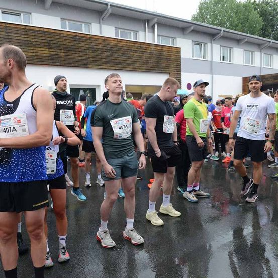 Marathonteilnehmer mit verschiedenen Startnummern und Kleidung stehen auf einer nassen Straße. Gebäude mit Fenstern und ein Schild sind im Hintergrund zu sehen.