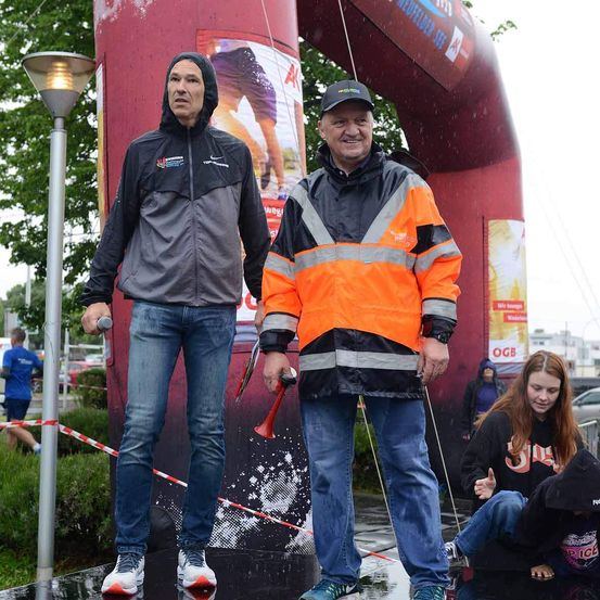 Zwei Männer stehen unter einem roten Bogen in einer regnerischen Außenumgebung. Ein Mann trägt eine reflektierende orangefarbene Jacke, der andere eine schwarze Jacke mit Kapuze. Hinter ihnen schaut ein junges Mädchen nach unten.