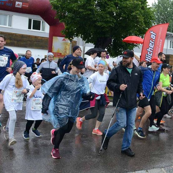 Ein Marathon ist im Gange mit mehreren Teilnehmern, darunter ein Mann mit einem Stock und eine Frau in einer Regenjacke. Die Veranstaltung findet wahrscheinlich bei regnerischem Wetter statt.