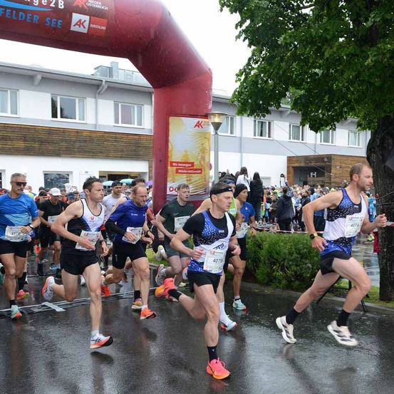Marathonläufer in nassen Kleidern laufen in einer Reihe vor einem Gebäude und einem Bogen mit Werbung.