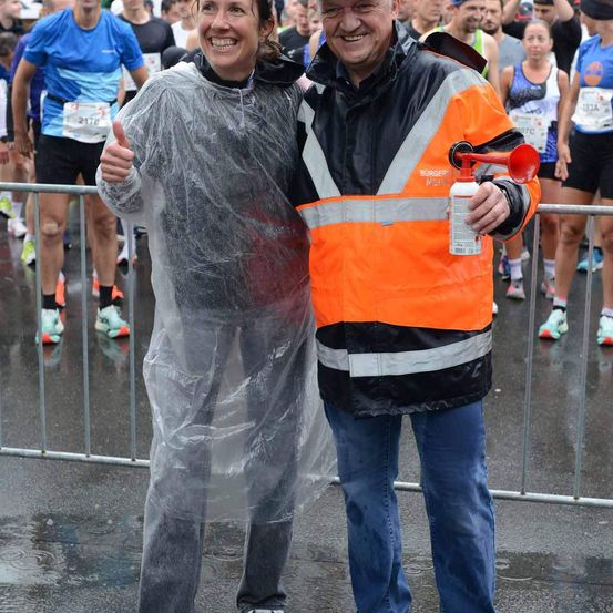 Eine Frau in einem Regenponcho und ein Mann in einem orangefarbenen Sicherheitsjackett stehen hinter einer Metallbarriere. Der Mann hält eine Hupe und lächelt die Frau an. Im Hintergrund laufen mehrere Menschen einen Marathon.