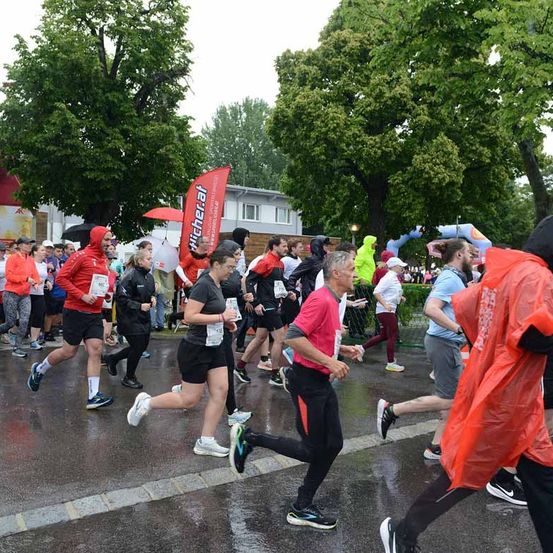 Eine Gruppe von Läufern nimmt an einem Marathon bei Regen teil. Sie sind in Regenkleidung gekleidet und einige halten Regenschirme. Bäume und ein Gebäude sind im Hintergrund zu sehen.