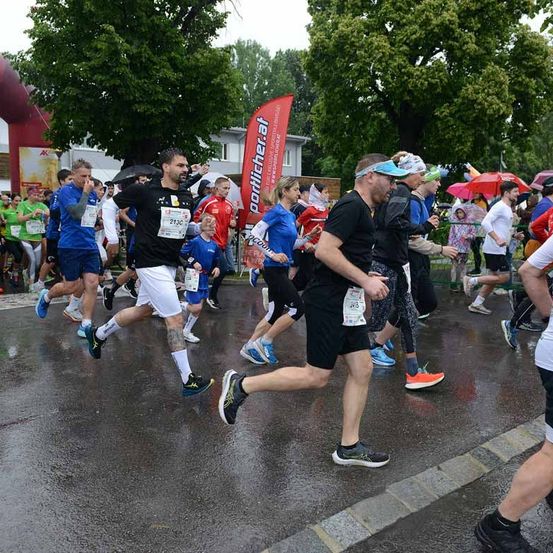 Eine Gruppe von Menschen läuft auf einer nassen Straße bei einem Marathon. Einige tragen Hüte und Sonnenbrillen. Ein Banner mit dem Wort porliberat steht hinter ihnen. Bäume und Gebäude sind im Hintergrund.