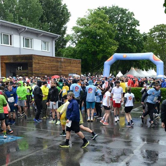 Eine Menschenmenge in Laufbekleidung versammelt sich auf einer nassen Straße, möglicherweise für einen Marathon. Sie warten auf den Start des Rennens. Ein Bogen ist in der Ferne.