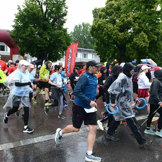 Läufer nehmen bei regnerischen Bedingungen an einem Marathon teil. Einige tragen Regenmäntel und Mützen, während andere Regenschirme haben. Bäume und Gebäude säumen den Kurs.