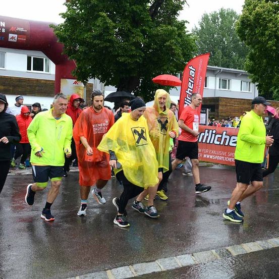 Eine Gruppe von Läufern in Regenmänteln läuft bei einem Marathon. Dahinter befindet sich ein Gebäude mit einem roten Banner und Bäumen.