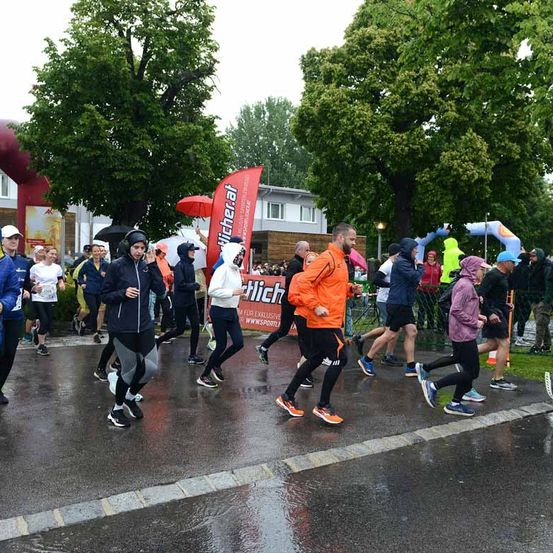 Eine Gruppe von Menschen läuft bei einem Marathon-Event. Sie tragen verschiedene Kleidungsstücke und Schuhe. Einige tragen Mützen und halten Regenschirme. Im Hintergrund sind Bäume und ein Gebäude zu sehen.