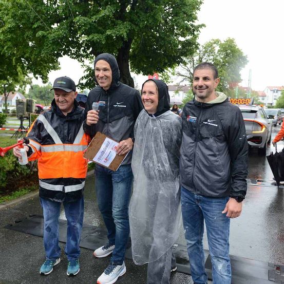 Vier Menschen stehen im Regen und lächeln für die Kamera. Ein Mann hält ein Klemmbrett und ein Mikrofon, ein anderer hat eine Pfeife, während zwei Frauen Regenmäntel tragen. Autos und Gebäude sind im Hintergrund.